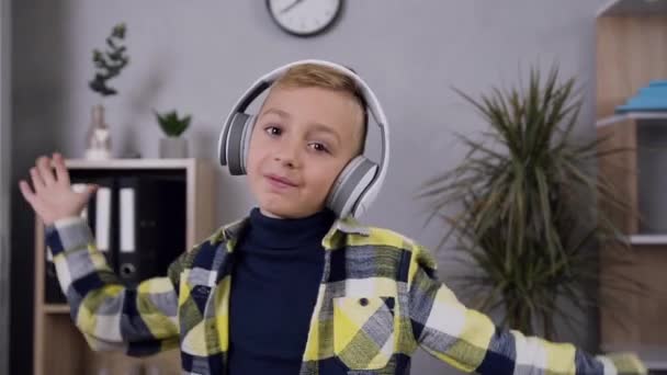 Vooraanzicht van lachende dansende tiener jongen in koptelefoon die poseren op camera in zijn kamer — Stockvideo