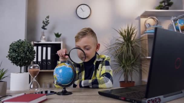 Attractive teen smiling boy looking at globe through the magnifying glass at home — Stock Video