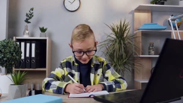 Vista frontal de niño moderno de 10 años de buen aspecto con gafas que anotan las notas en el cuaderno desde el ordenador — Vídeo de stock