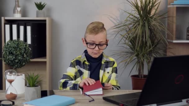 Handsome voorzichtig 10-jarige jongen met bril zitten aan de tafel en het schrijven van de notities in het dagboek met behulp van de computer — Stockvideo