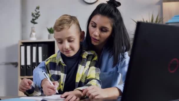 Όμορφη νεαρή σκούρα μαλλιά γυναίκα γράφει κάτι στο σημειωματάριο μαζί με το χαμογελαστό έφηβο αγόρι της στο τραπέζι στο σπίτι — Αρχείο Βίντεο