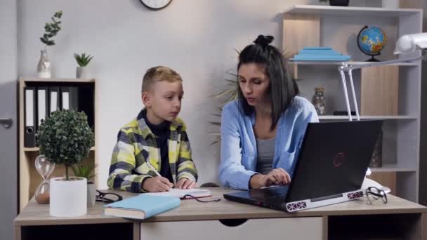 Gut aussehende ernsthafte brünette Frau, die am Computer arbeitet, während ihr hübscher, ausgeglichener Teenager-Sohn in ihrer Nähe Schulunterricht gibt — Stockvideo