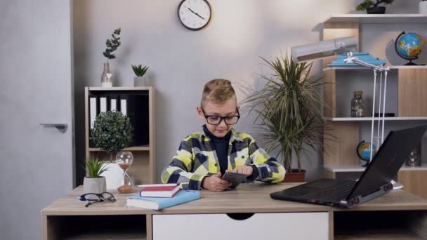 Bonito satisfeito inteligente adolescente menino em óculos sentado na mesa em casa e usando telefone e computador — Vídeo de Stock