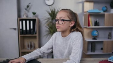 Sevimli, modern odaklı, 15 yaşında, gözlüklü bir kız odasında bilgisayarla çalışıyor.