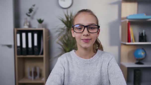 Likable friendly smiling 15-aged girl in glasses posing on camera on the background of her room — Stock Video