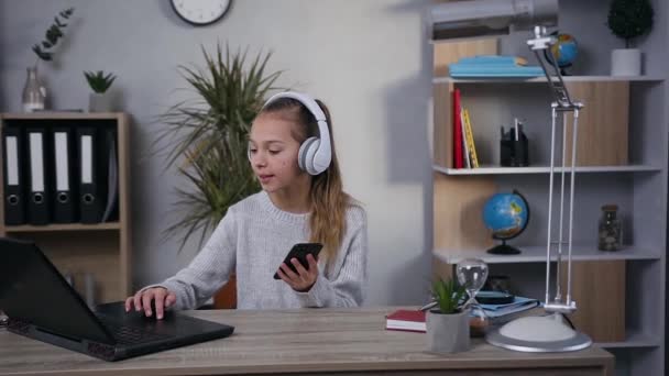 Niedliche übermütige moderne Teenie-Mädchen mit Kopfhörern genießen die coole Musik am Tisch in ihrem Zimmer — Stockvideo