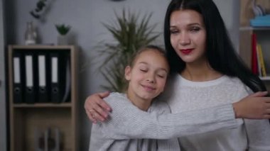 Mutlu, şık bir anne ve genç kız birbirlerine sarılıyorlar ve evde kameraya bakıyorlar.