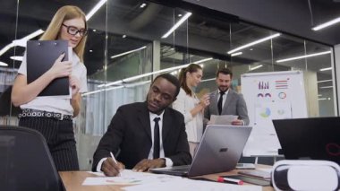 Attractive satisfied african american businessman sitting at boardroom table and signing the documents on the background of talking business colleagues