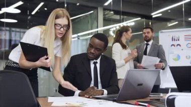 Attractive high-spirited bearded businessman talking with his experienced female business colleague while another multiracial business colleagues revisioning important documents