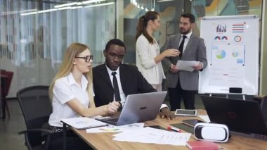 Close view of good-looking experienced 30-35s multiracial business people which working at computer while two another business colleagues talking near whiteboard with charts