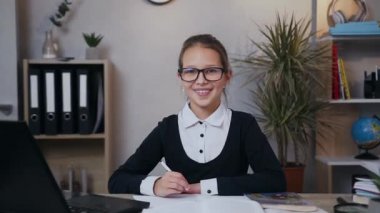 Gülümseyen mutlu üniformalı okul kızının ön görüntüsü. Ödevinin başında oturup kameraya bakarken bir fikir düşünüyor.