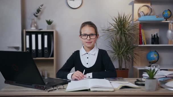 Přední pohled na dobře vypadající veselá teen dívka v brýlích, které se dívá na kameru se šťastným úsměvem, zatímco dělá doma její školní úkol — Stock video