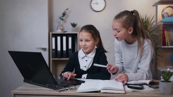 Frontansicht eines ziemlich ernsthaften 15-jährigen Mädchens, das ihrer jüngeren Schwester hilft, ihre Hausaufgaben mit dem Computer zu machen — Stockvideo