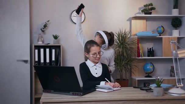 Vista frontal de la divertida chica adolescente bonita en auriculares que bailan bajo música inflamatoria, mientras que otra chica sonriente escribiendo notas en el cuaderno — Vídeos de Stock