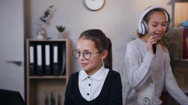 Hermosa colegiala sonriente de 10 años en gafas mostrando algo interesante a su hermana mayor que baila bajo música moderna en auriculares — Vídeos de Stock