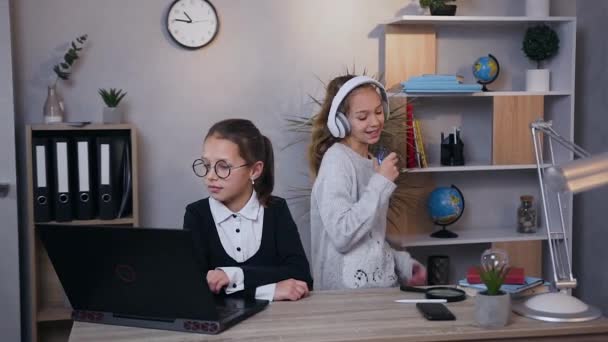 Цілком приваблива дівчина-підліток танцює під запальною музикою в навушниках і ще одна молодша спокійна дівчина, що працює на комп'ютері — стокове відео