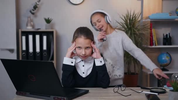 Porträt eines wütenden attraktiven Teenie-Mädchens, das die Schläfen vor Schmerzen massiert, weil ihre ältere lustige Schwester mit Kopfhörern sie mit Gesang und Tanz unter Musik irritiert — Stockvideo