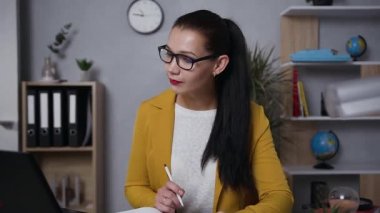 Balanced good-looking modern brunette businesswoman in glasses making notes on report with charts in front of laptop screen in home office
