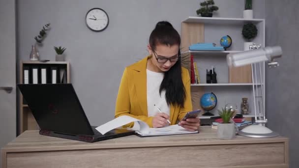 Közelkép a meglehetősen koncentrált szorgalmas fiatal üzletasszonyról szemüvegben, aki a kezében tartja a telefont, miközben grafikonos jelentéssel dolgozik. — Stock videók