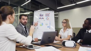 Handsome confident bearded businessman- coach explaining to his business multiracial group the value of graph using flip chart presentation