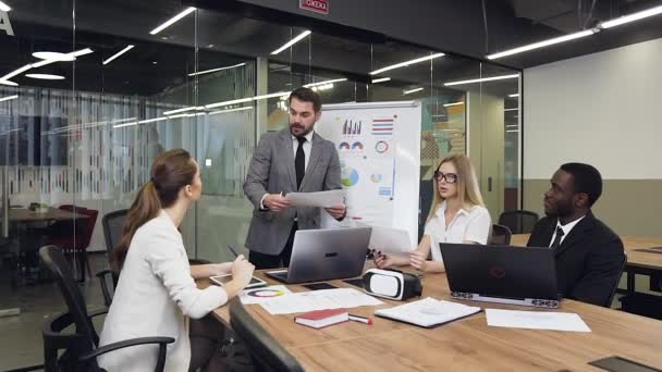 Atractivo hombre de negocios barbudo experimentado en traje de pie cerca de la presentación del rotafolio y hablando con su equipo de negocios multiétnicos industriosos — Vídeo de stock