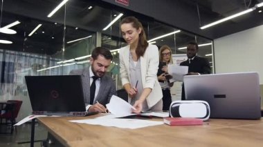 Close up of attractive satisfied bearded businessman which signing the documents in meeting room which giving him pretty young female worker on the background of talking partners