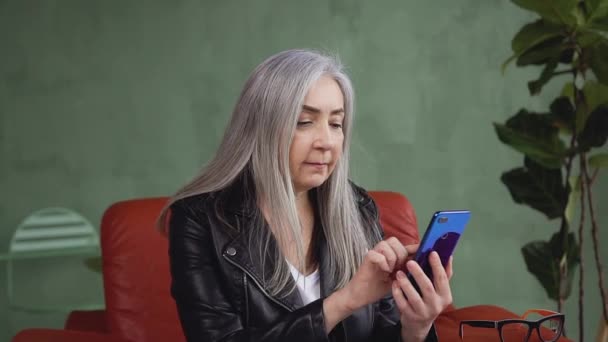 Щаслива усміхнена зріла жінка з довгим сірим волоссям сидить у зручному кріслі і використовує свій телефон — стокове відео