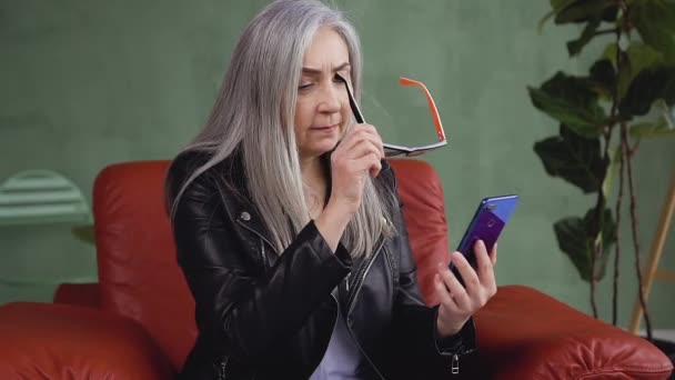 Front view of attractive gray hair stylish woman which putting on her eyeglasses while browsing the phone apps — Stock video