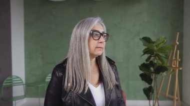 Front view of good-looking stylish 60-aged modern woman in glasses with long gray hair which posing on camera with lovely smile