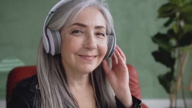 Beautiful smiling friendly retired woman with gray hair listening music in earphones and looking at camera with joyful face