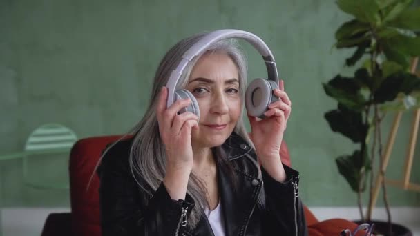 Вид спереди привлекательной улыбающейся фривольно пожилой женщины с длинными седыми волосами, которая надевает наушники и смотрит в камеру — стоковое видео