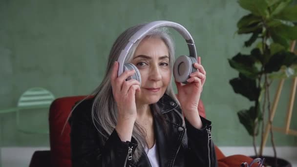 Front view of appealing smiling freindly senior woman with long gray hair which putting on headphones and looking at camera — ストック動画