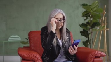 Portrait of charming modern 60-aged woman in glasses which using her smartphone while sitting in comfortable chair