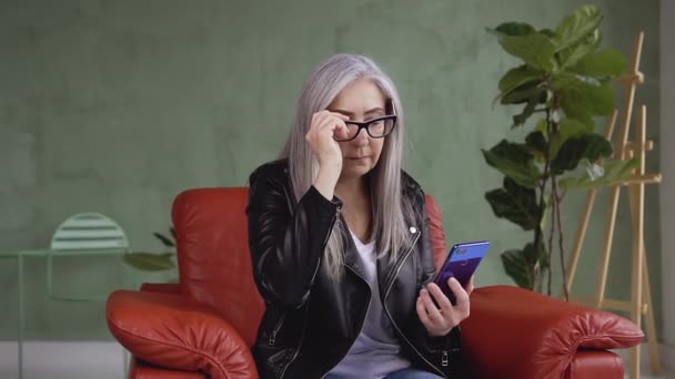 Retrato de una encantadora mujer moderna de 60 años en gafas que usa su teléfono inteligente mientras está sentada en una silla cómoda — Vídeos de Stock