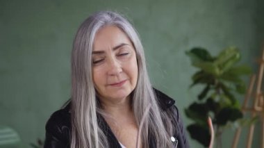 Smiling attractive confident gray haired woman in stylish jacket posing on camera while putting on her eyeglasses