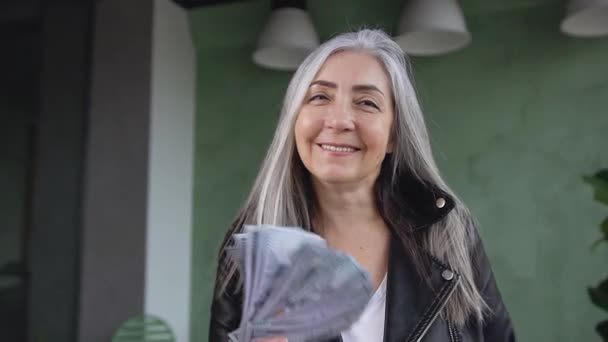 Вид спереди на счастливую богатую привлекательную пожилую модную женщину, которая держит в руках банкноты в долларах и позирует на камеру с счастливой улыбкой — стоковое видео
