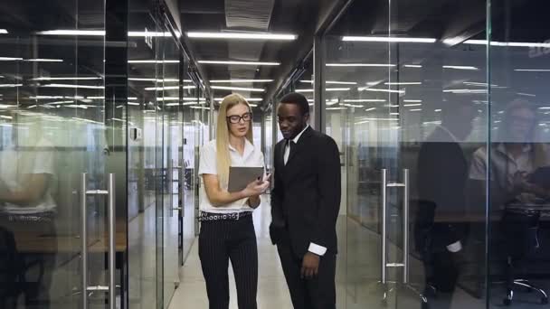 Вид спереду на двох задоволених багаторасових бізнесменів, які стоять в сучасному залі компанії і обговорюють дані з i-pad — стокове відео