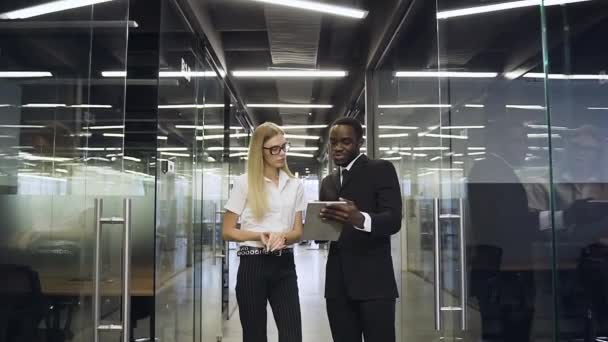 Good-looking serious 30s multiethnic business people standing in glass office hall and discussing aboult business project using tablet pc — Stock video