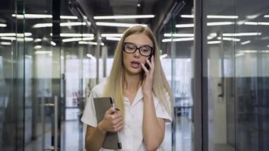 Front view of splendid smiling high-skilled businesswoman in glasses which talking on phone while walking through the office corridor