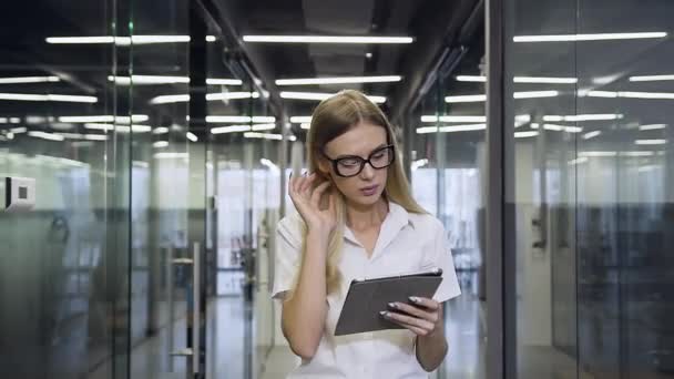 Серьезная бизнесвумен в очках ходит по современному офисному залу и пользуется планшетным компьютером — стоковое видео