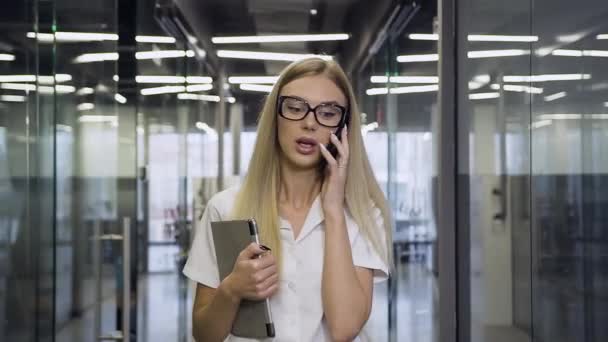 Vue de face de splendide femme d'affaires souriante et hautement qualifiée dans des lunettes qui parlent au téléphone tout en marchant dans le couloir du bureau — Video