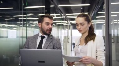 Attractive serious young business people discussing datas using computer and reports while standing in company hall with glass offices