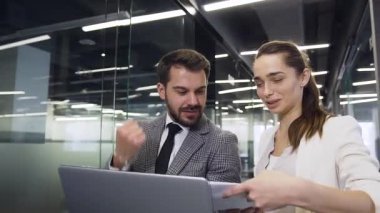 Good-looking satisfied modern young business colleagues standing with laptop in beautifully designeted with glass office hall