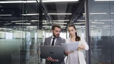 Attractive satisfied professional young businessman and businesswoman walking through the long glass office hall and talking about deatails of joint work