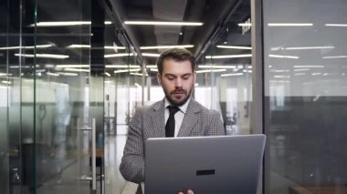 Good-looking concentrated young bearded businessman with computer walking through the modern company hall