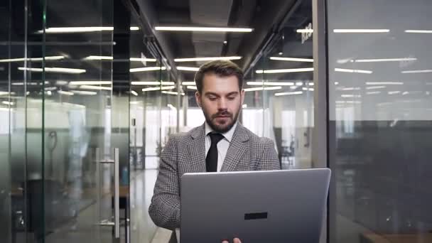 Красивый концентрированный молодой бородатый бизнесмен с компьютером, прогуливающийся по современному залу компании — стоковое видео