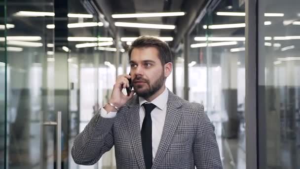Привлекательный успешный современный бородатый бизнесмен, прогуливаясь по офисному залу и разговаривая по телефону — стоковое видео