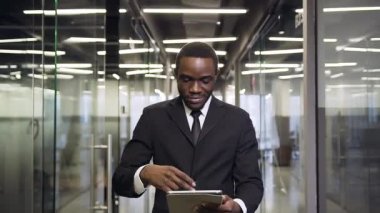 Good-looking serious dark-skinned businessman walking to his workplace through office corridor, using tablet pc and looking at camera