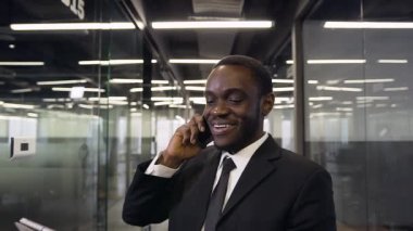Attractive smiling professional black-skinned office manager standing in long company corridor and talking on phone