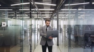 Close view of attractive confident bearded young businessman in suit which holding in his hands computer and posing on camera in modern company hall